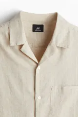 Camisa de manga corta color beige claro, confeccionada en mezcla de lino y algodón. Tiene cuello abierto, tapeta lisa, bolsillo superior abierto, canesú en la espalda y bajo recto con aberturas laterales.