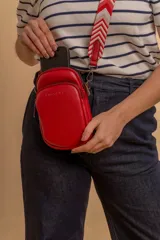 Bandolera pequeña tipo 'phone bag' de símil cuero color rosado pálido, con cierre de cremallera y correa ancha de tela con diseño de chevron en tonos claros.