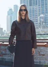 Chaqueta corta marrón de efecto piel, con cuello camisero, cierre frontal con botones, dos bolsillos de parche con solapa en la parte delantera y forro interior.