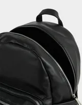 Mochila urbana negra de cuerina con bolsillo frontal con cierre y charm colgante de cadena con dije de corazón y candado. Tiene correas regulables, manija superior, cierre superior, doble bolsillo frontal (uno con charm metálico) y bolsillos laterales.