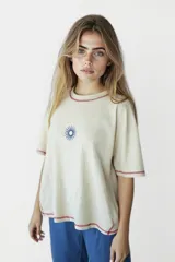 Remera beige de corte holgado con cuello redondo y mangas cortas. Presenta un pequeño estampado de un sol azul en el centro del pecho y costuras en color rojo en cuello, mangas y ruedo.