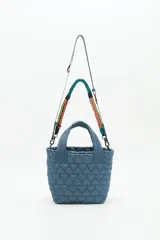 Cartera tote azul de nylon matelassé con diseño de corazones. Tiene manijas cortas y dos correas desmontables, una de cuero sintético y otra de tela trenzada multicolor. Cuenta con cierre superior y dos bolsillos internos.