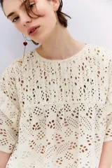 Blusa color crudo con bordado calado tipo broderie, de corte holgado, escote redondo, manga corta con volados en la sisa y detalle de alforzas en el canesú.