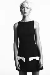 Vestido corto negro con cuello redondo y sin mangas. Presenta dos lazos blancos en la parte inferior delantera y aberturas frontales en el ruedo. Cierre trasero con cremallera metálica.