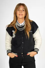 Campera tipo universitaria negra con mangas blancas, cierre con botones a presión y detalles de rayas en puños y cintura. Presenta bordados con letras y números en el frente y la espalda.