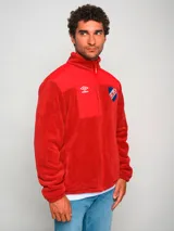 Campera polar roja con cierre frontal, cuello alto y bolsillos laterales. Presenta un panel liso en la parte superior del pecho y un bolsillo con cierre en el lado izquierdo. Incluye el logo de Umbro bordado en el pecho izquierdo y el escudo del Club Nacional de Football bordado en el pecho derecho.