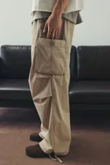 Pantalón cargo de algodón color beige, de corte holgado y tiro medio. Presenta cintura elástica con cordón ajustable, bolsillos laterales y grandes bolsillos cargo con solapa en las piernas. Los bajos son ajustables con cordón oculto.