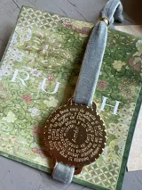 Pulsera dorada con medalla redonda con la oración del Ave María grabada en español, y cinta de terciopelo gris.