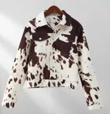 Chaqueta con estampado de vaca en tonos marrón y blanco, con cuello camisero, cierre frontal con botones metálicos, bolsillos de parche en el pecho y corte recto.