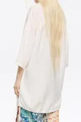 Camisa larga color blanco, de corte oversize y escote con solapas. Tiene manga corta recta y ancha, cierre con botonadura frontal y bajo ajustable con cordones de tejido.