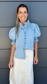 Camisa celeste de denim con cuello mao y mangas cortas abullonadas con elástico. Presenta detalle de punto smock en cuello y mangas, y cierre frontal con botones.