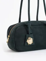 Cartera rectangular de estructura firme confeccionada en cuero descarne vacuno color marrón. Presenta manijas fijas, cierre superior y un charm metálico dorado en forma de vaquita de San Antonio colgando de un tag lateral. Incluye el logo de la marca grabado en relieve en el frente.