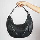Cartera al hombro negra de cuero sintético con forma de media luna. Tiene cierre superior y dos bolsillos frontales con cierre.