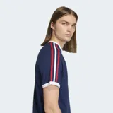 Remera Adidas de manga corta, color azul marino, con cuello redondo blanco. Presenta las tres franjas características de la marca en las mangas en colores rojo, blanco y azul, y el logo del trébol en dorado en el pecho.