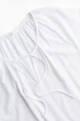 Blusa blanca de tejido liviano con mangas raglán cortas. Modelo de corte holgado con escote redondo levemente fruncido, abertura en V y tiras finas para atar adelante.