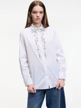 Camisa blanca a rayas verticales finas, con cuello clásico y cierre frontal con botones. Presenta apliques de pedrería en el cuello y en la tapeta de los botones.