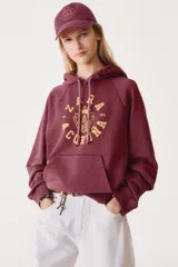 Sudadera color borgoña con capucha ajustable con cordones, manga larga y estampado dorado en el frente con el texto "A Coruña" y un escudo.