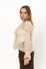 Camisa color crema con estampado floral en tonos naranja, con cuello en V y volados frontales. Mangas largas abullonadas con puños elásticos.