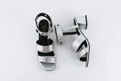 Sandalias plateadas con dos tiras horizontales sobre el pie y una tira vertical que se une a una pulsera con hebilla en el tobillo. La pulsera tiene ojales y tachas combinadas. El taco es cuadrado y de 5 cm.