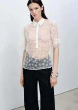 Camisa blanca semitransparente con bordado de flores, cuello camisero, manga corta y cierre frontal con botones.
