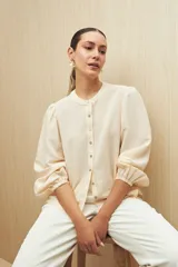 Camisa color crema a rayas verticales finas, de cuello mao, mangas 3/4 abullonadas con elástico en los puños y cierre frontal con botones de nácar.