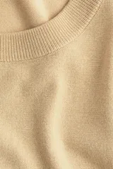 Buzo corto color beige de punto fino, con cuello redondo, hombros caídos y mangas largas.