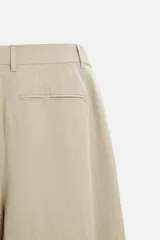 Pantalón color beige de corte ancho, con pliegues frontales, bolsillos laterales y traseros de doble vivo.