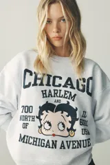 Sudadera gris melange de cuello redondo y manga larga con puños elásticos. Estampado frontal con texto "Chicago Harlem" y dibujo de Betty Boop.