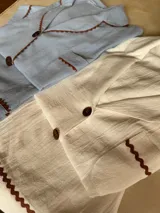 Pijama de algodón color blanco, compuesto por una camisa de manga corta abotonada con cuello solapa y un bolsillo en el pecho, y un short con elástico en la cintura. Ambas prendas tienen un detalle de picot marrón en los bordes.