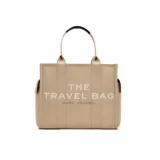 Bolso tote beige de lona con asas superiores de cuero negro y bolsillo lateral. Estampado frontal con el texto "THE TRAVEL BAG MARC JACOBS".