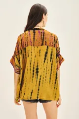 Camisa de manga corta con cuello mao y calce amplio, confeccionada en viscosa con teñido tie-dye en tonos mostaza, negro y violeta. Presenta bordados florales con mostacillas, lentejuelas y canutillos en tonos turquesa y dorado en la parte delantera.