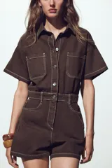 Mono corto color verde oliva, de denim con cuello solapa y manga corta. Tiene bolsillos delanteros y traseros, cintura elastizada en la espalda y cierre frontal con cremallera y botones metálicos.