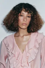 Camisa color rosa pálido confeccionada en tejido ligero con lyocell. Presenta escote en V, cierre frontal con botones, mangas largas abullonadas con puños elásticos y detalle de volantes en el frente.