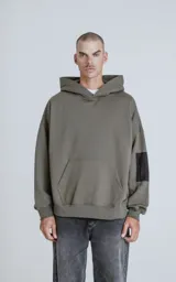 Hoodie color verde militar de rústico 100% algodón, con silueta oversize, capucha forrada y bolsillo tipo bomber en la manga con cierre. Presenta bolsillo canguro frontal.