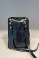 Bolso pequeño tipo bandolera o cartera de mano, confeccionado en cuero vacuno color taupe/marrón claro, con detalles en material metalizado plateado. Presenta un bordado de flor de lis en el frente y cierre superior. Incluye correa fina desmontable.