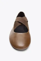 Zapato tipo bailarina plana color marrón, confeccionado en piel. Presenta tiras elásticas cruzadas en la parte delantera y acabado en punta redonda.