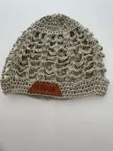 Gorro tipo beanie tejido a mano en hilo de algodón, color beige jaspeado, con diseño de malla calada y etiqueta de cuero marrón con el nombre 'Soriame' grabado.