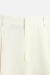 Pantalón color crema de lino, corte amplio, con pliegues frontales, bolsillos laterales y traseros de ojal. Cierre frontal con cremallera y gancho metálico oculto.