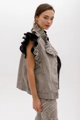 Chaleco de denim color topo o gris claro lavado, con volados estructurados en los hombros y a lo largo del frente. Es de corte recto, sin mangas, con cierre frontal de botones metálicos color cobre y bolsillos laterales.