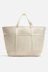 Bolso tipo shopper color crema, fabricado en tejido de algodón. Tiene doble asa de mano, compartimento principal con cierre de cremallera, seis bolsillos planos interiores, funda interior para dispositivos electrónicos de hasta 13 pulgadas con cierre de cremallera, bolsillo plano frontal y bolsillos planos laterales.