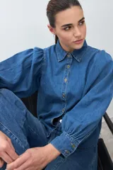 Camisa de jean de color azul medio, con cuello camisero, botones dorados y mangas largas abullonadas con puños ajustados.