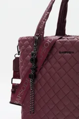 Bolso tote grande acolchado color vino tinto, con costuras en patrón de rombos. Posee dos asas de mano y una correa ajustable y desmontable con texto repetido. Incluye un charm colgante con letras negras.
