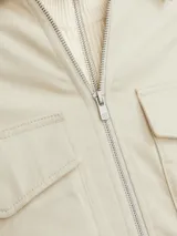 Chaqueta color crema, de manga larga, con cuello camisero y cierre frontal con cremallera metálica. Tiene dos bolsillos de parche con solapa en el pecho.