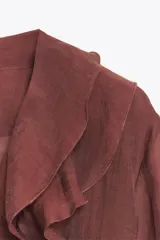 Camisa color crudo confeccionada en tejido con lyocell. Presenta escote en pico, mangas largas abullonadas con elástico en los puños, volantes frontales y cierre frontal con botones.