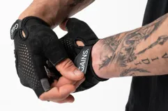 Guantes de entrenamiento sin dedos, color negro y rosa. La parte superior es de malla transpirable rosa con bordes grises reflectantes. La palma es negra con un patrón de agarre hexagonal gris. Cuentan con una tira de velcro en la muñeca con la inscripción "GAINS.".