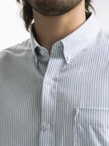 Camisa celeste y blanca a rayas verticales, de manga larga y corte clásico. Tiene cuello abotonado y bolsillo en el pecho.