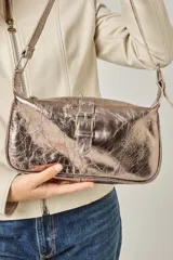 Bandolera de gamuza color gris topo, con correa ajustable con hebilla y aplique de hebilla metálica en el frente. Cuenta con cierre superior, bolsillo trasero con cierre y forro interior de algodón con bolsillos.