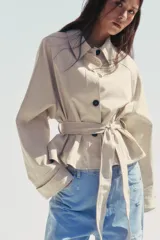 Trench corto color crema con cuello solapa, manga larga con trabilla, bolsillos de doble vivo, forro interior y cinturón lazada en el mismo tejido.