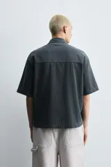 Camisa de denim de algodón, color beige claro, con corte relajado, cuello bowling y manga corta. Presenta cierre frontal con botones a presión y un pequeño bordado a tono en el pecho.