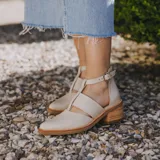 Zapato Kira de cuero color beige, con tira central y pulsera con hebilla al tobillo. Tiene taco de madera de 5 cm y cierre trasero con cremallera.